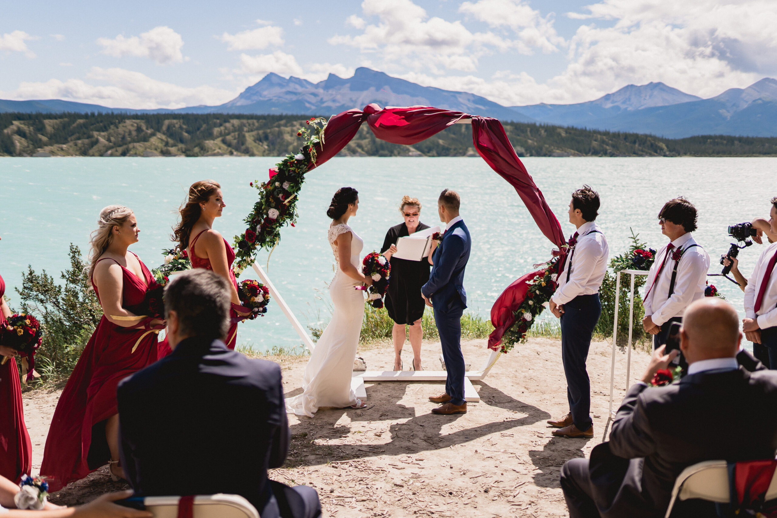 Lakefront wedding ceremony with red wedding ceremony arch and bridesmaids in red gowns