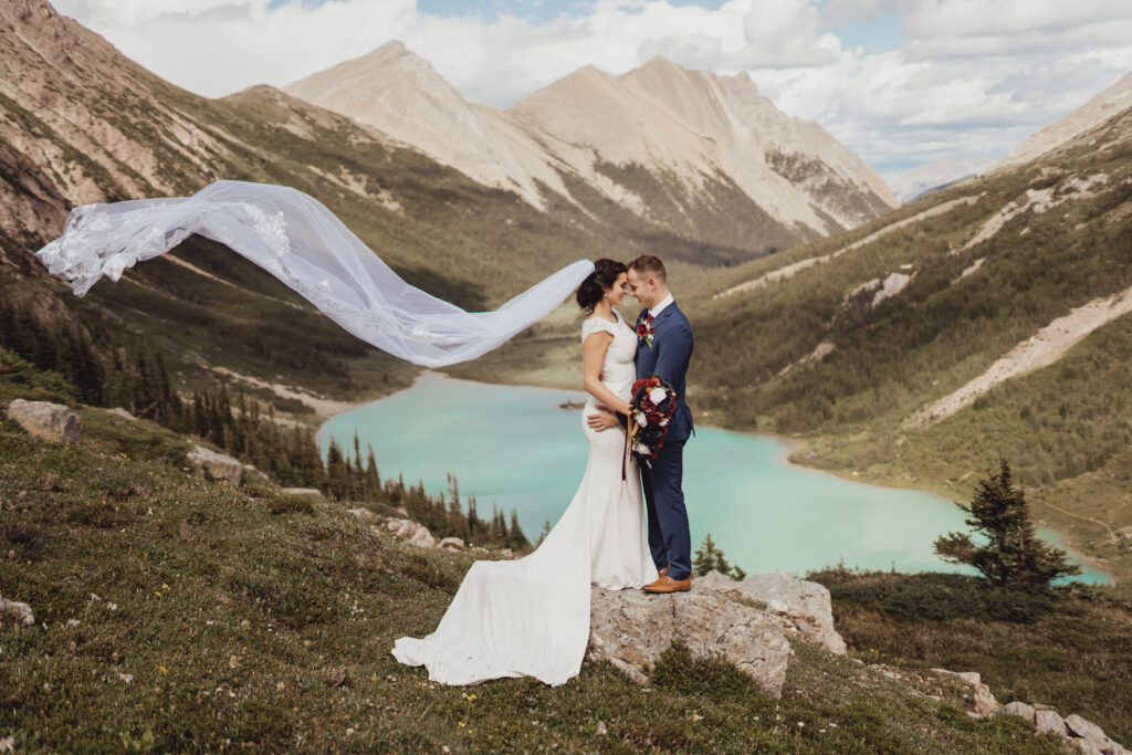 Bride and groom kissing in front of pristing blue lake, cathedral veil blowing in wind, Rocky Mountain landscape