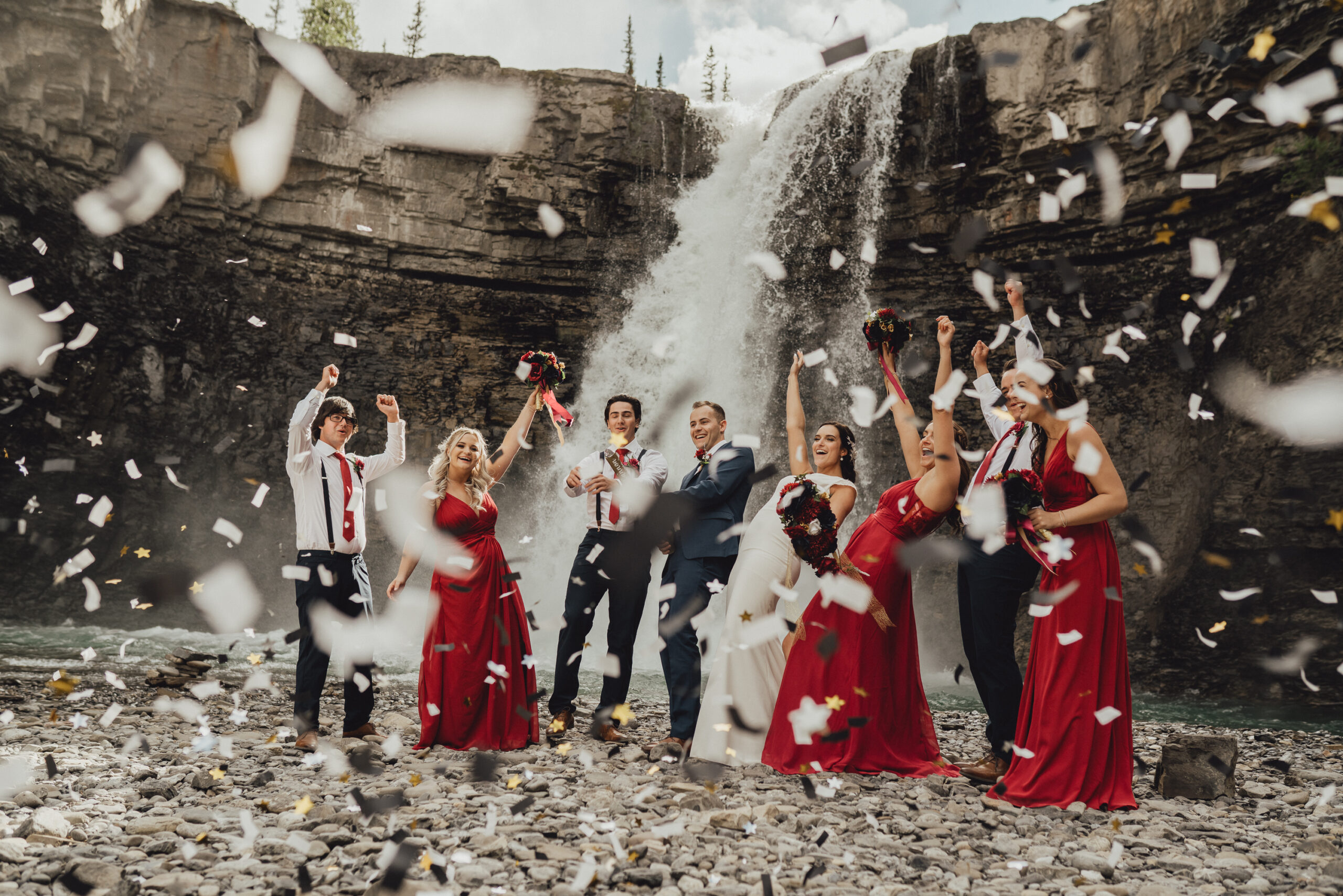 Bride in classic wedding gown with bridesmaids in red bridesmaids gowns celebrating in front of waterfall