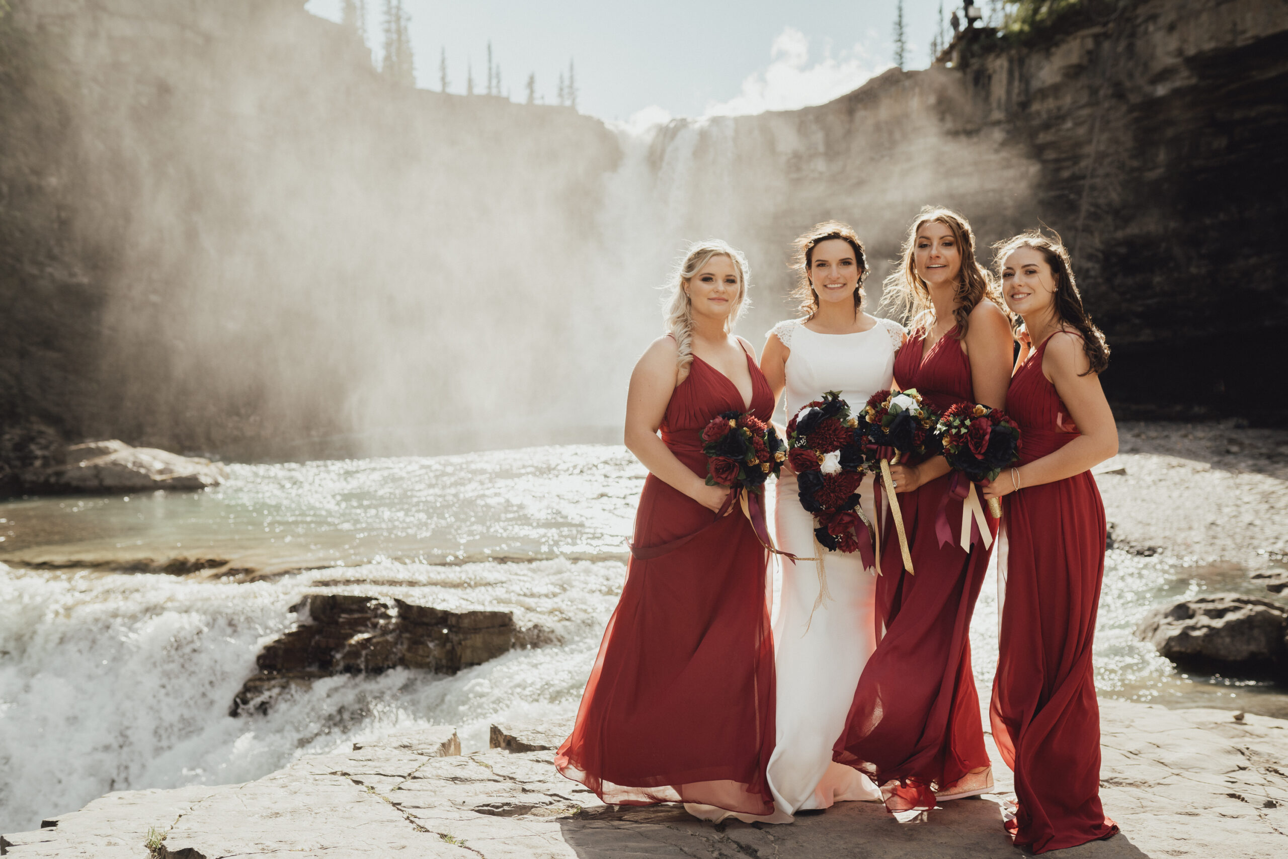 Bride in classic wedding gown with bridesmaids in red bridesmaids gowns standing in front of waterfall