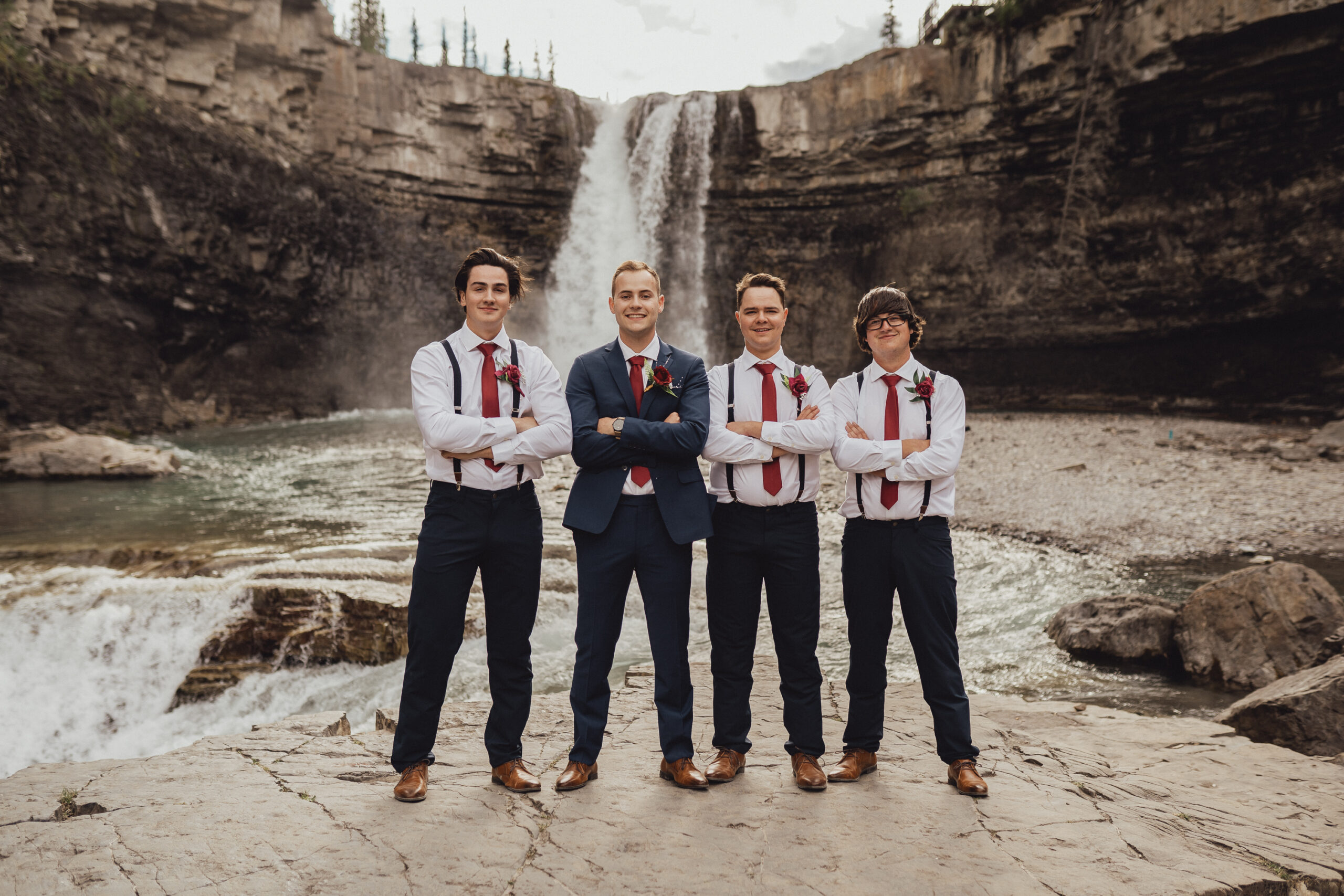Groom in cobalt blue wedding suit and groomsmen with red suspenders standing in front of waterfall