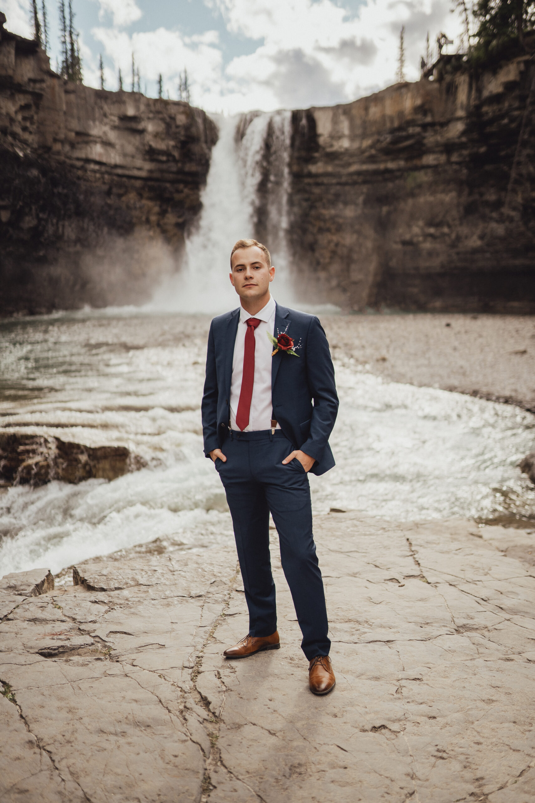 Groom in cobalt blue wedding suit and red tie standing in front of waterfall