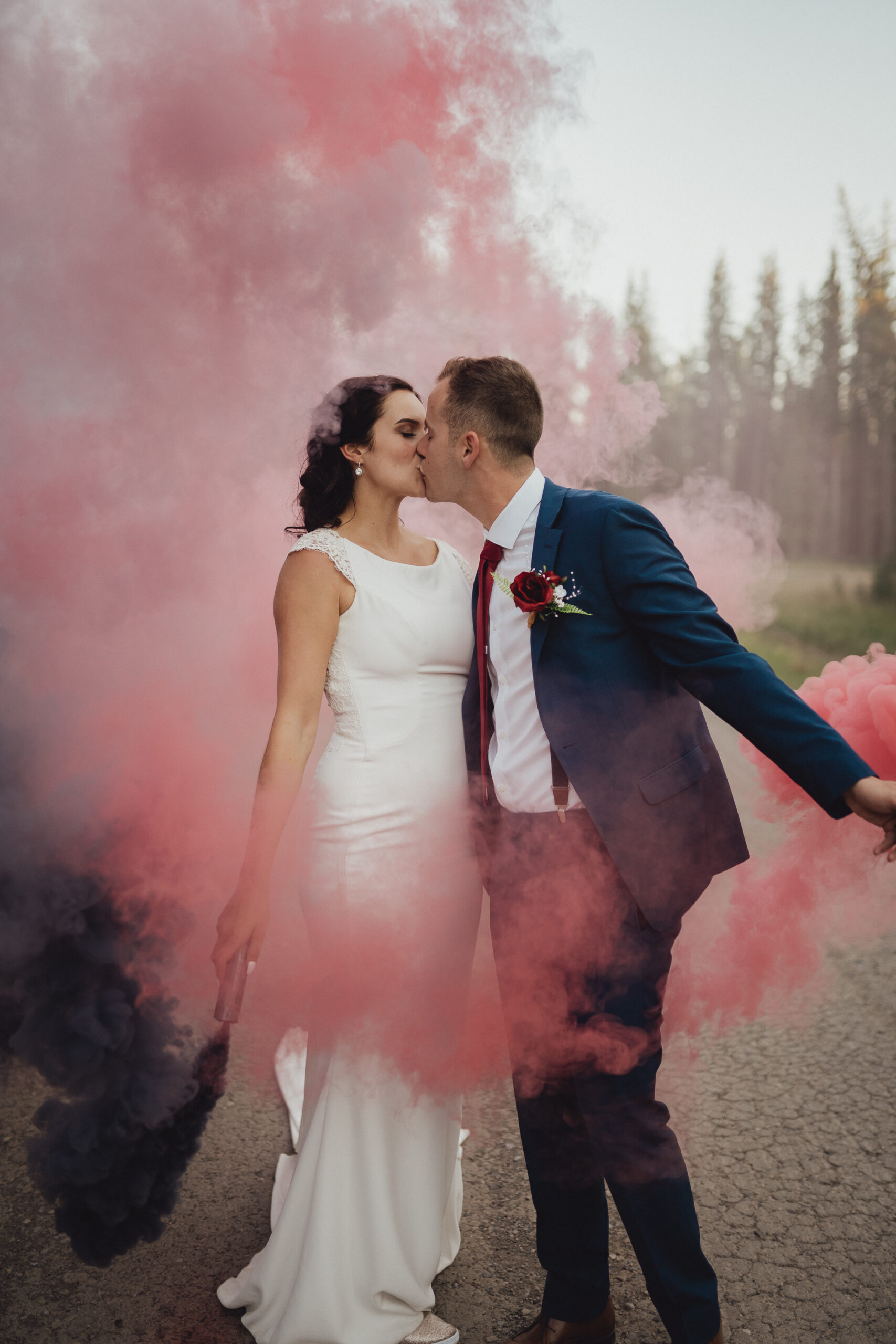 Bride in high neck gown and groom in cobalt blue suit kissing in the middle of coloured red smoke
