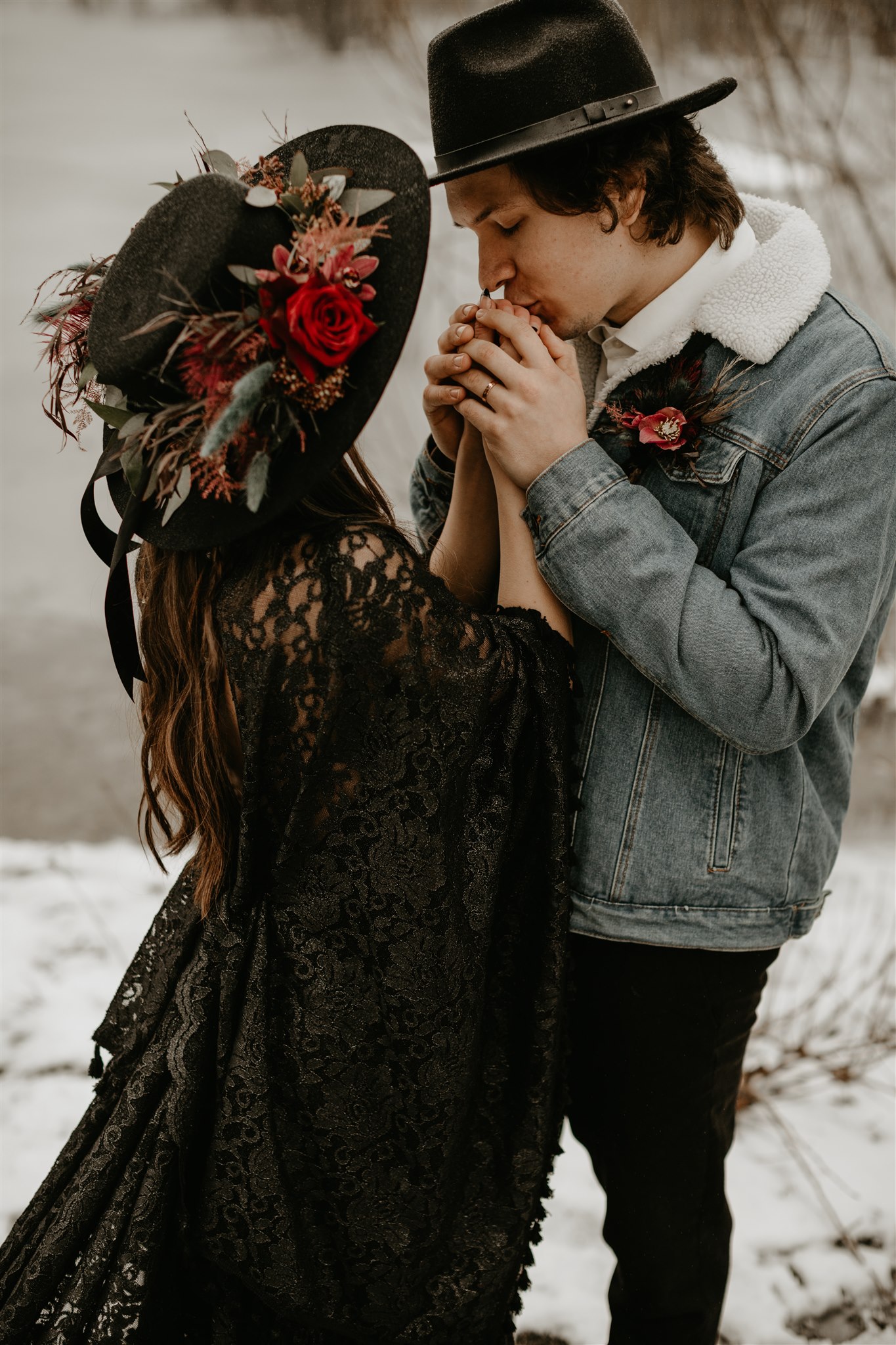 Boho bride in black wedding dress with black wedding hat with red flower garland and boho groom in jean jacket