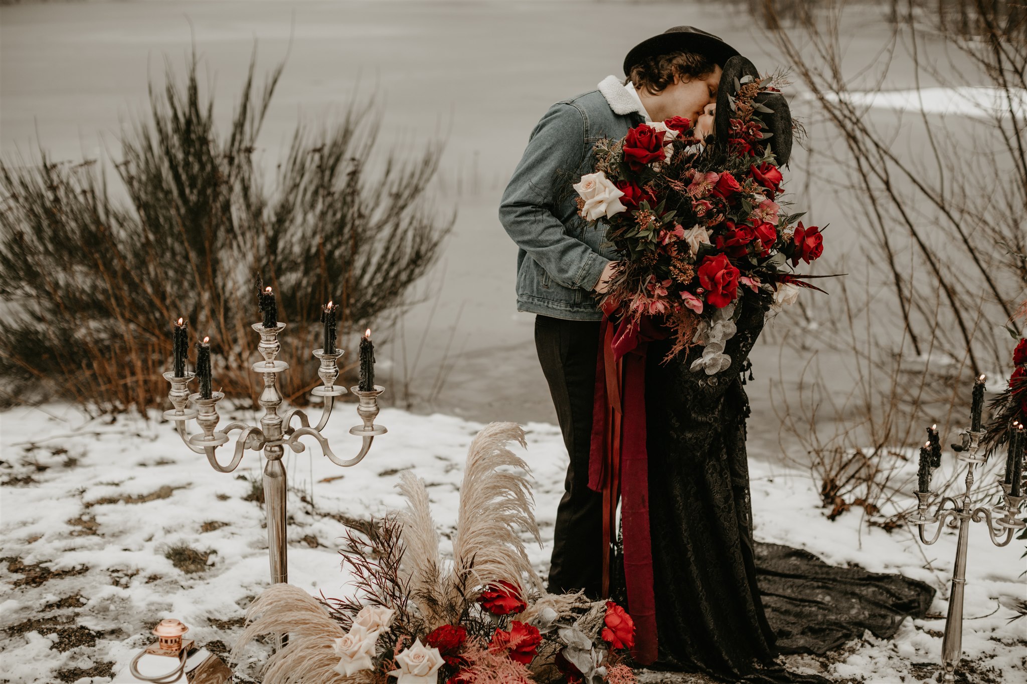 Boho winter bride in black wedding dress and black wedding hat kissing groom in jean jacket in front of river
