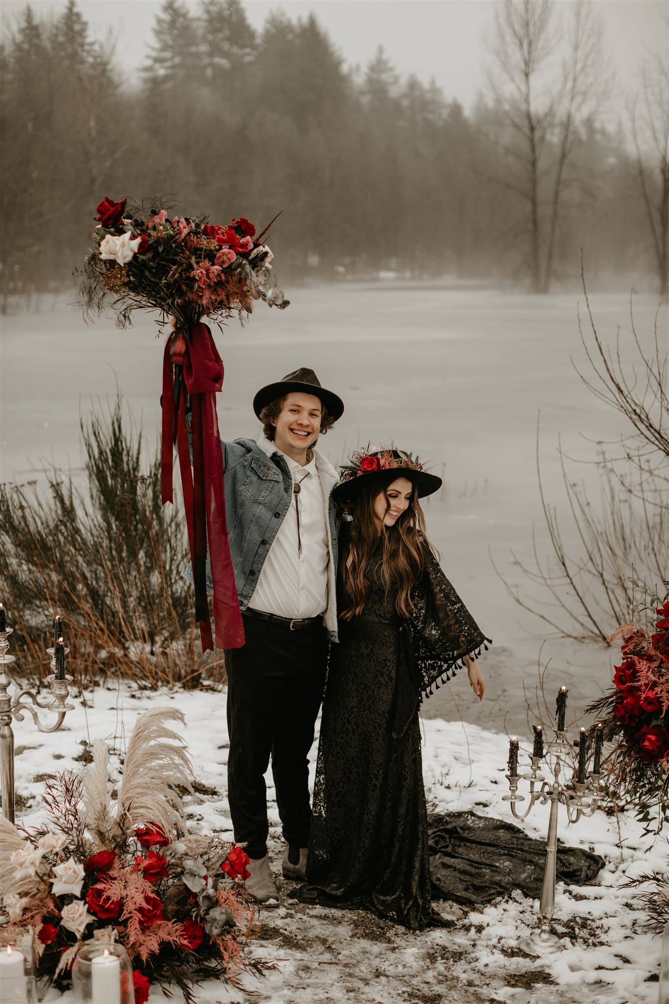 Boho bride in black wedding dress with black wedding hair and groom holding red wedding bouquet above his head on a snowy river bank