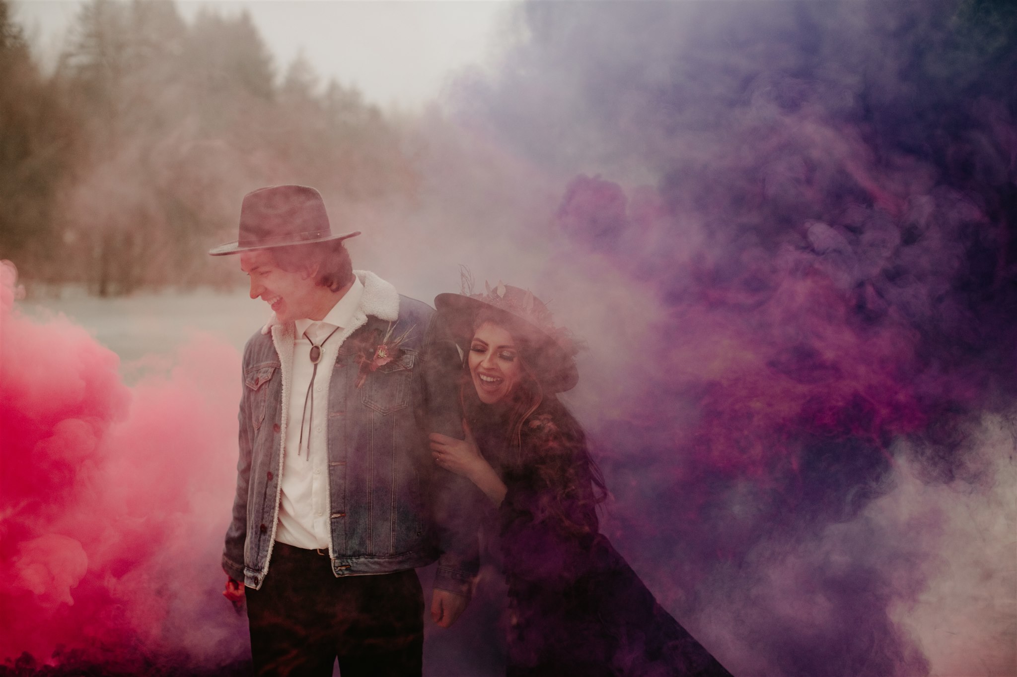 Boho chic bride in furry jacket and groom in jean jacket on snowy river bank with coloured purple and red smoke