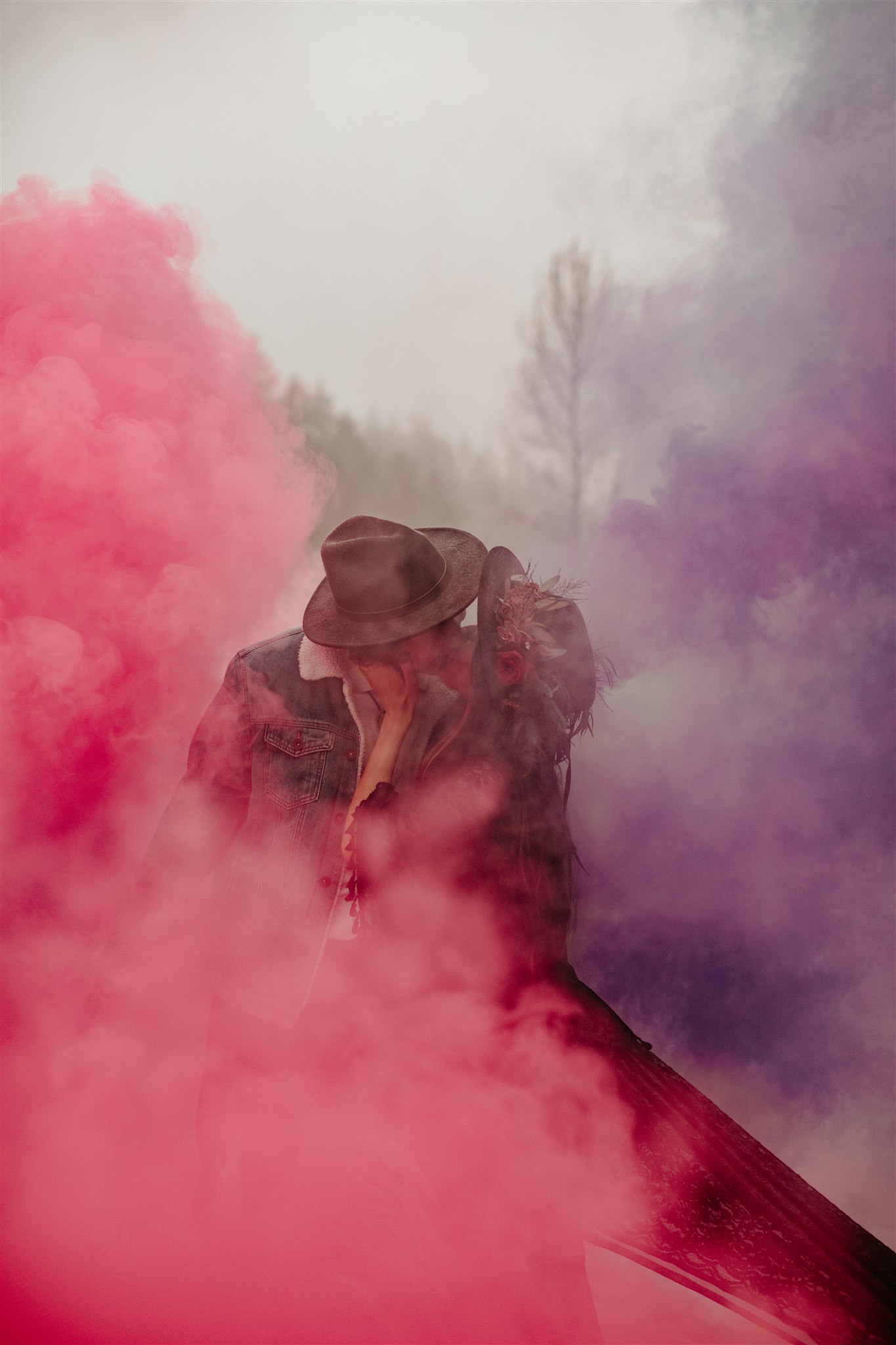 Boho chic bride in furry jacket and groom in jean jacket on snowy river bank with coloured purple and red smoke