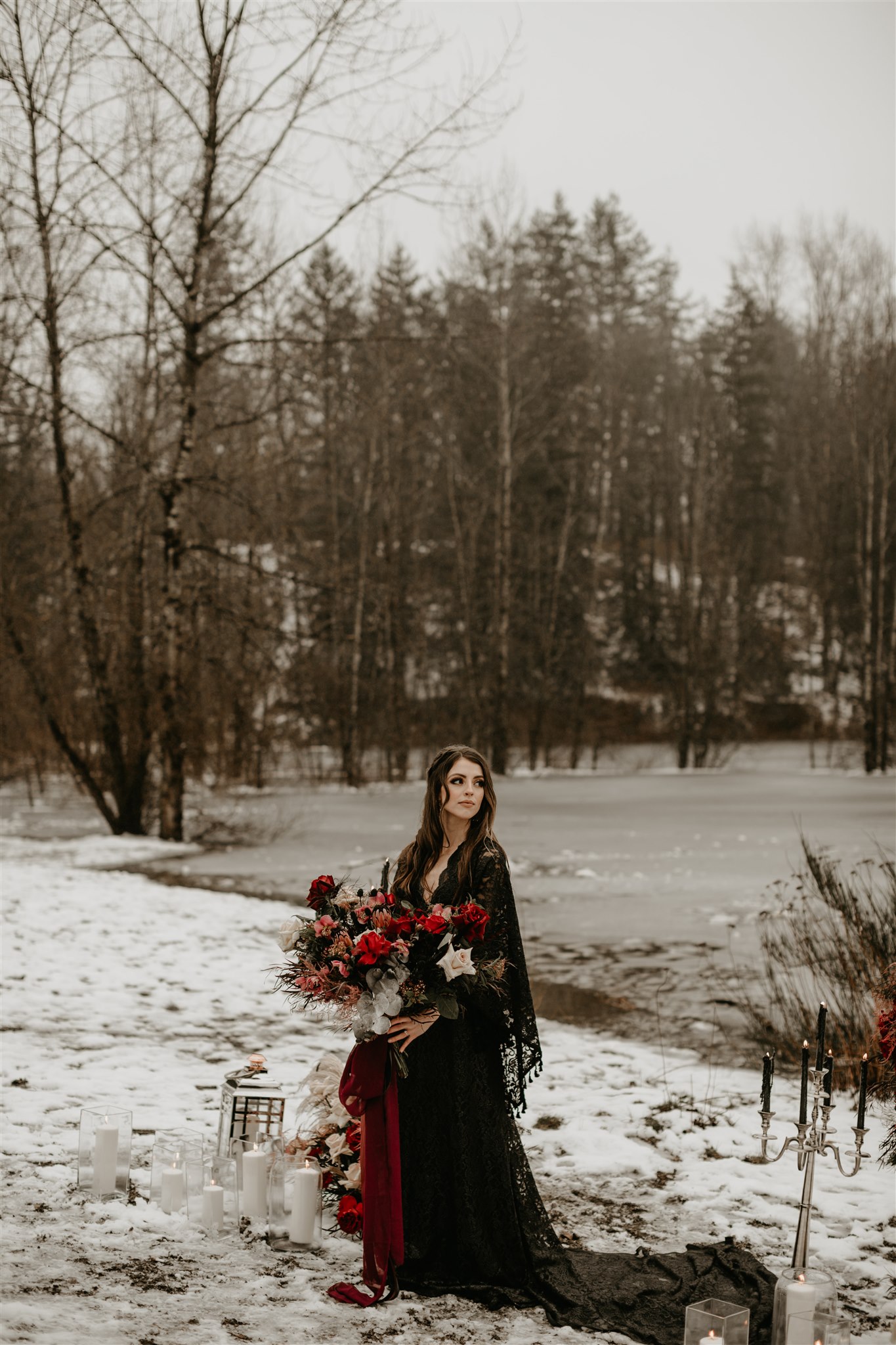 Boho winter bride in black wedding dress and black wedding hat on snowy river bank