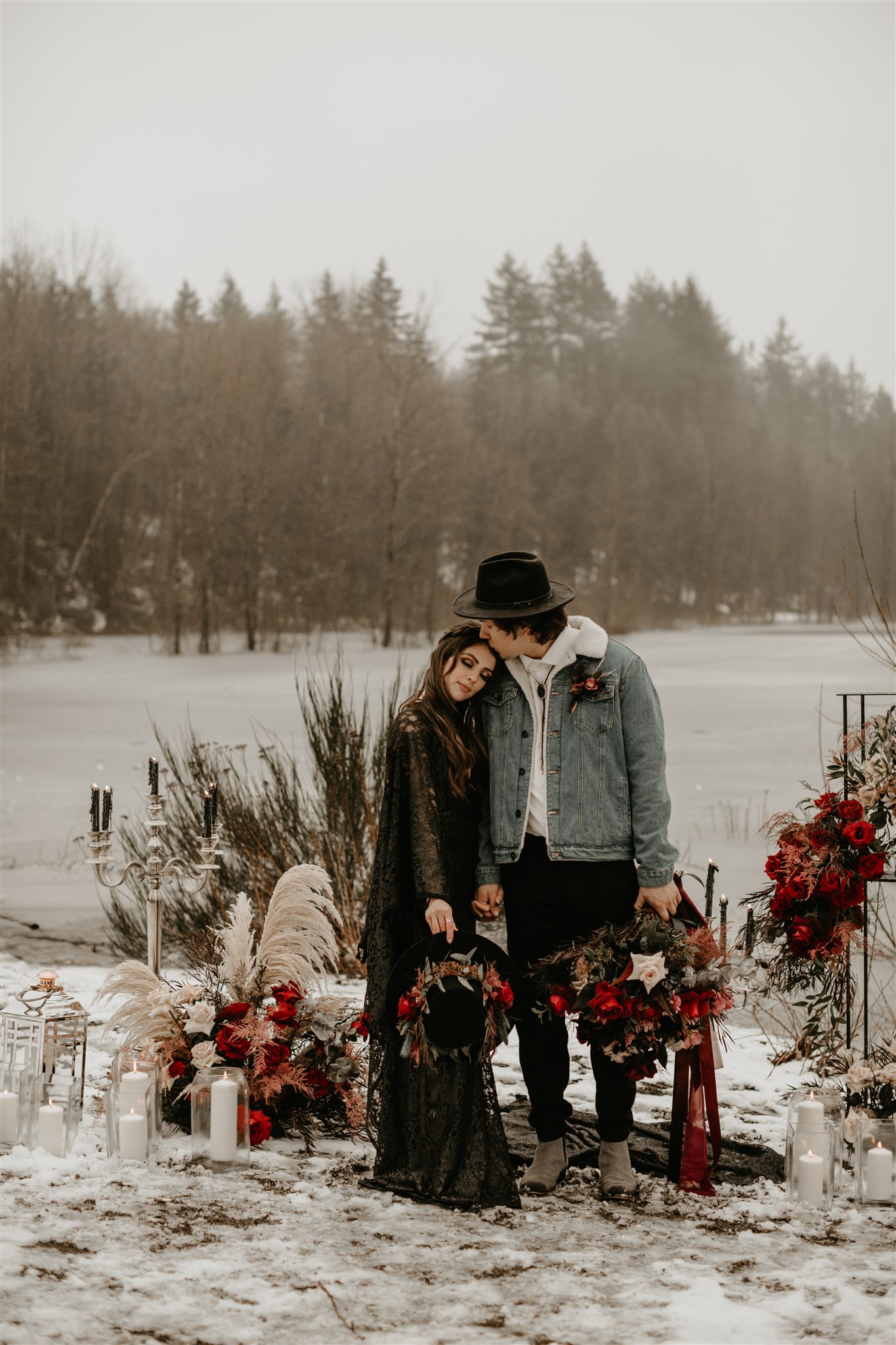 Boho chic bride in furry jacket and groom in jean jacket on snowy river bank in front of boho ceremony set up