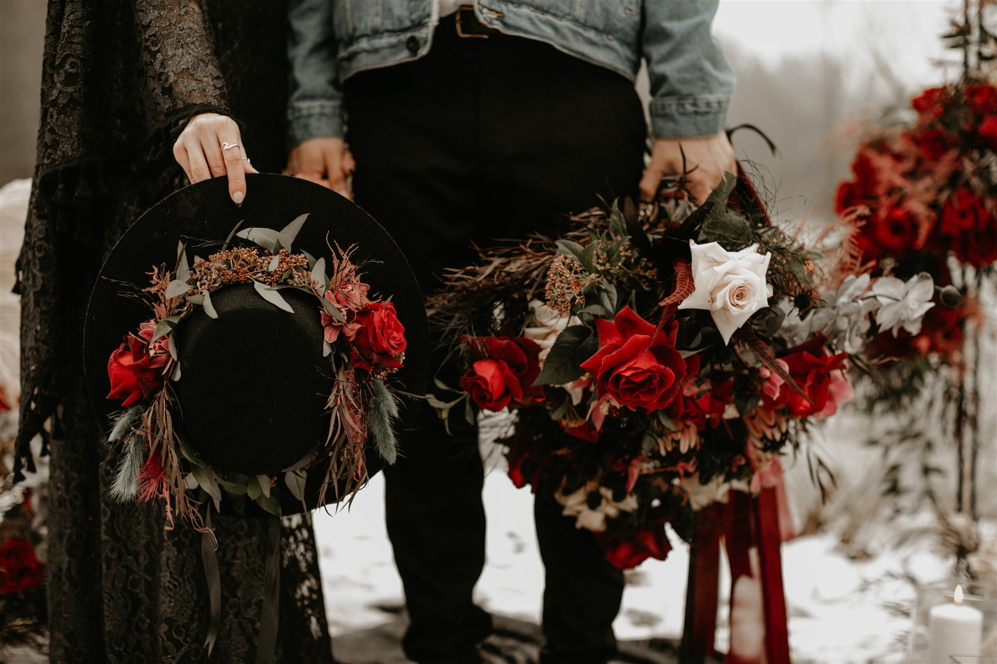 Boho chic bride in furry jacket and groom in jean jacket on snowy river bank holding boho chic red wedding bouquets