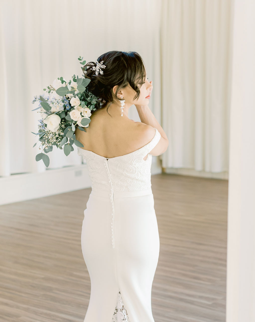 Dark hair bride with low chignon holding light blue and white bouquet over her shoulder wearing a low back wedding dress