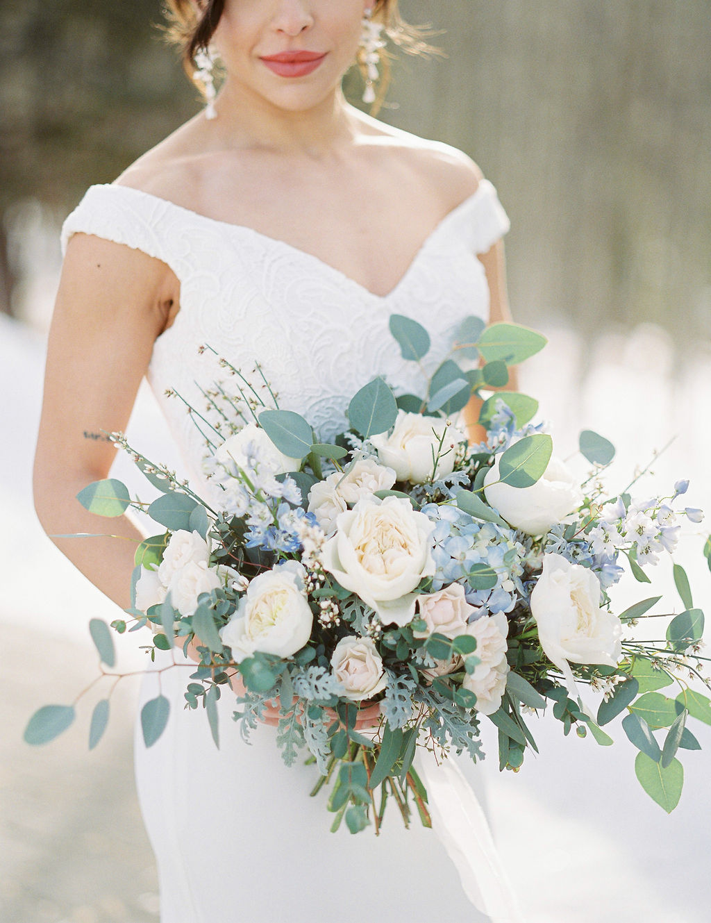 Classic bride with bold lipstick and chandelier earrings in an off the shoulder gown with a sweetheart neckline holding a light blue and white bouquet