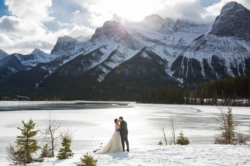 Bride and groom kissing on snowy river bank with Rocky Mountains in the backdrop