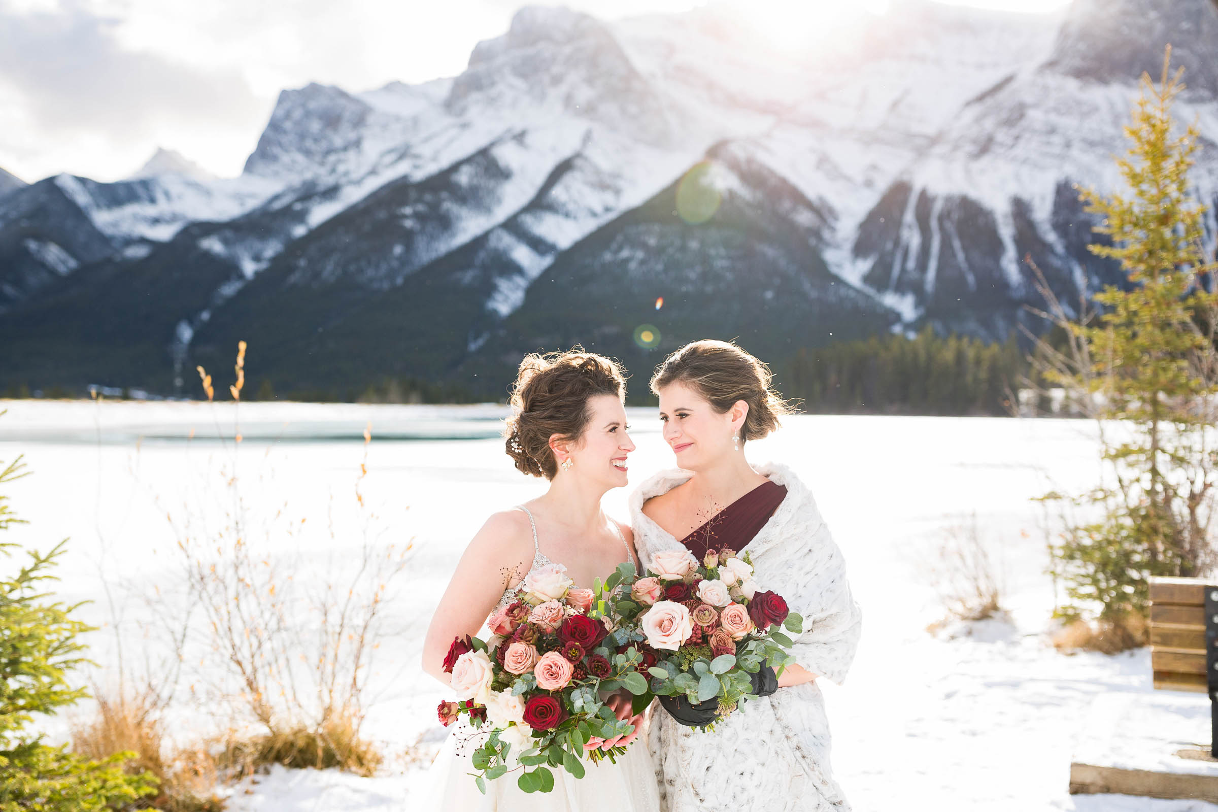 Classic bride and maid of honour smile at each other holding wedding bouquets of red, blush and white roses