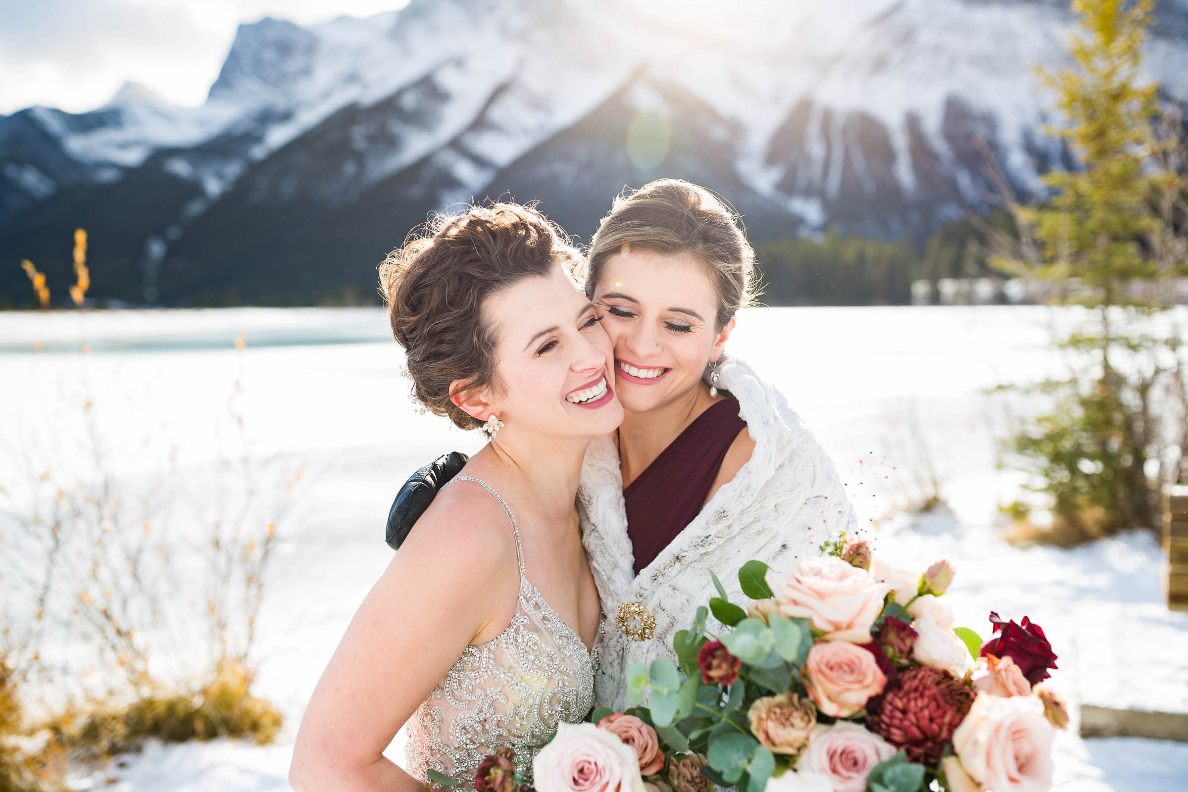 Bride and maid of honour laughing and hugging each other holding wedding bouquets of red, blush and white roses