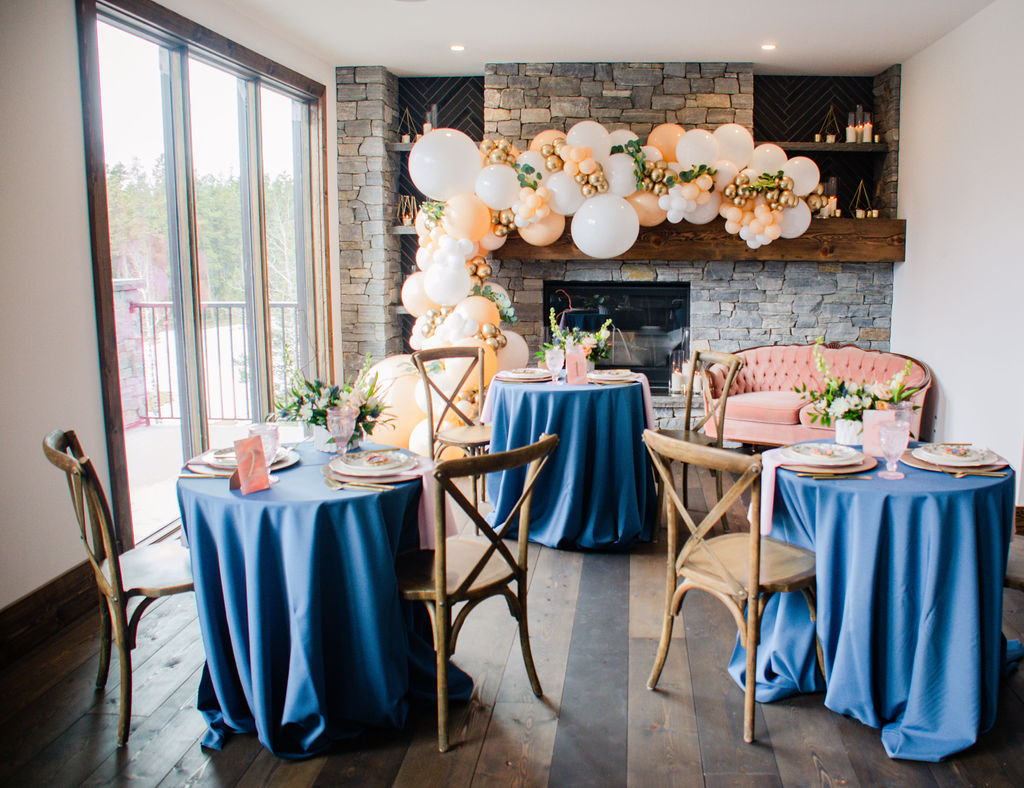 Small tables with blue tableclothes, peach and white balloons against a stone fireplace