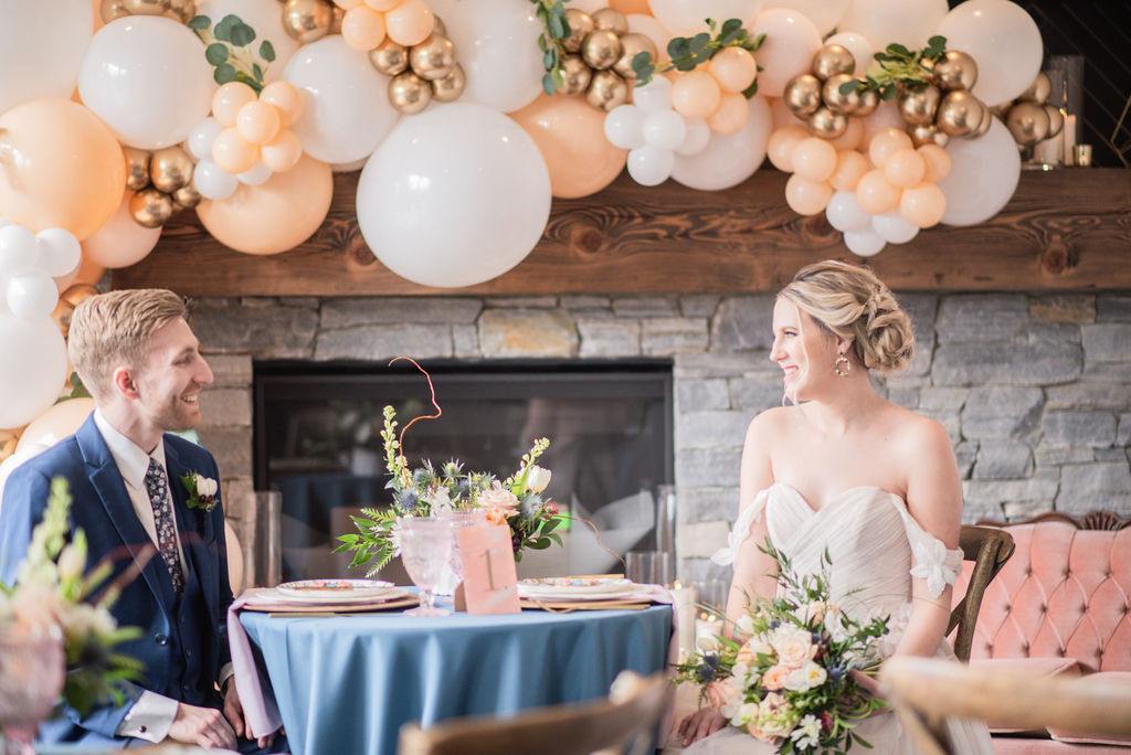 Bride and groom sitting at sweetheart table with blue tablecloth and peach and white balloons in the background