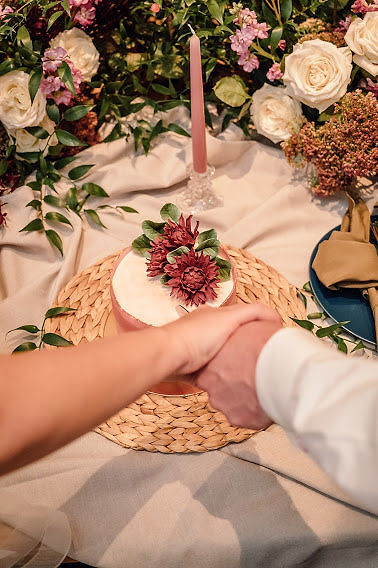 Bride and groom holding hands infront of small blush wedding elopement cake with blush water color details and burgundy flowers