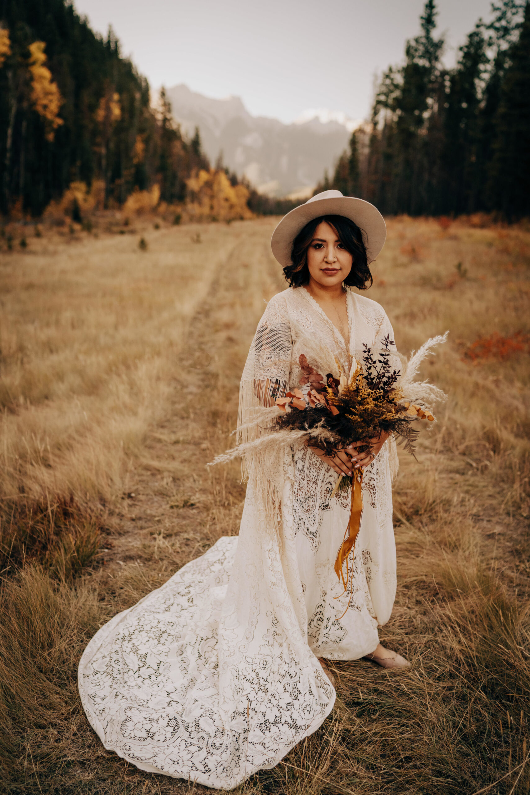 Boho bride in maternity lace wedding gown with hat holding bouquet of flowers in neutrals and browns