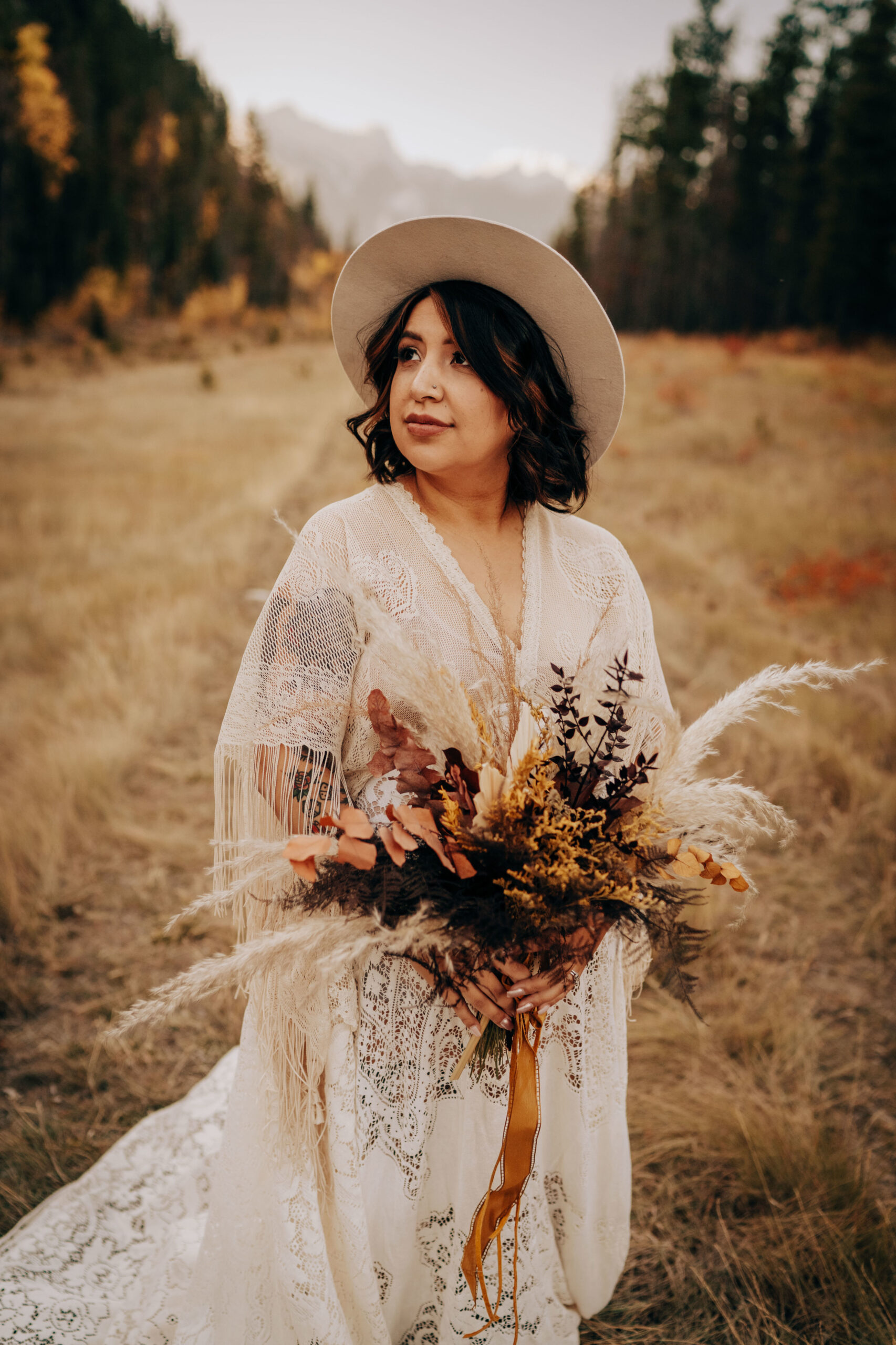 Boho bride in maternity lace boho wedding gown and hat standing in a valley bolding bouquet of neutrals and brown with