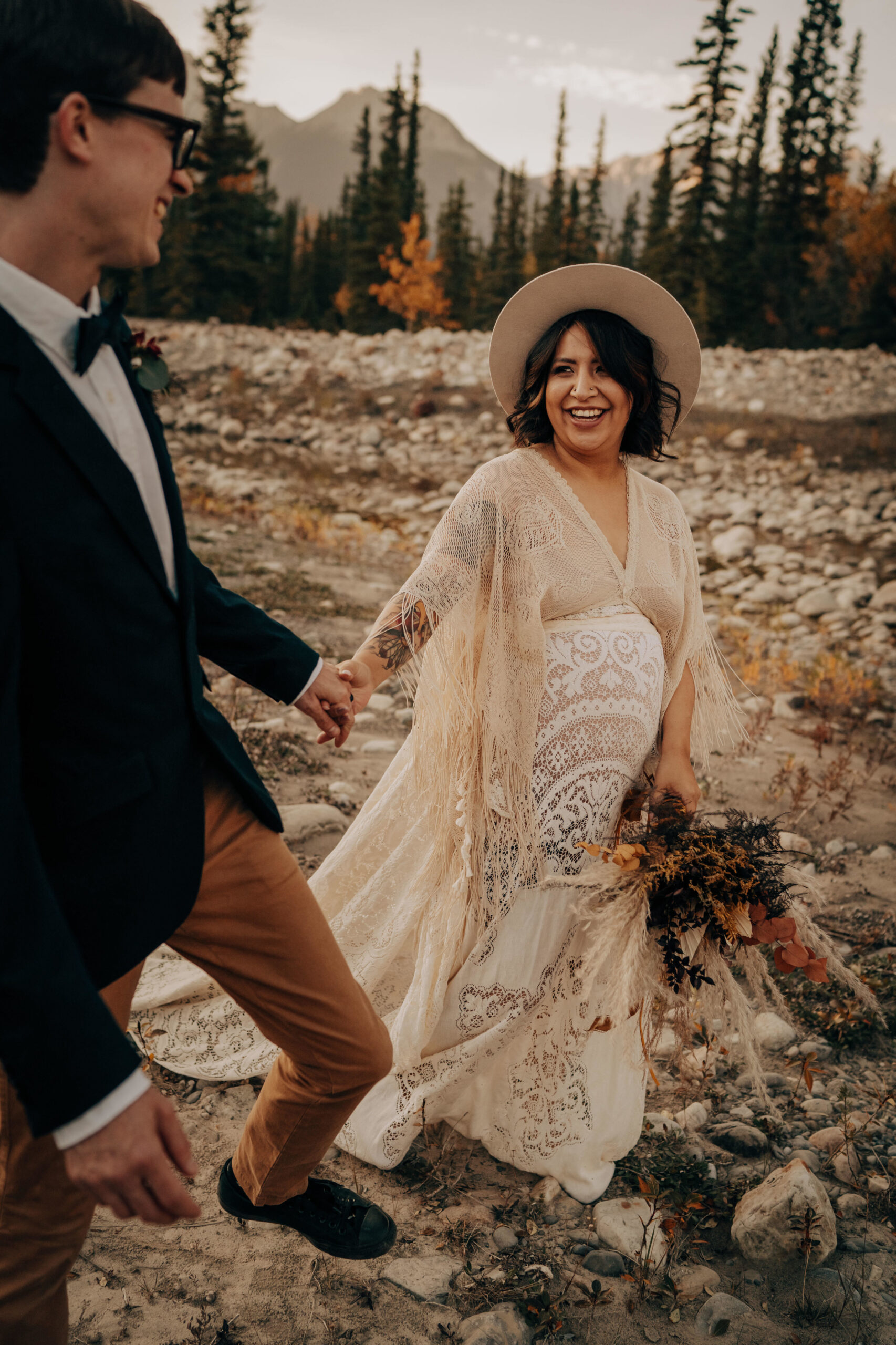 Boho bride in lace boho maternity wedding gown and boho groom standing in valley, holding hands and looking at each other
