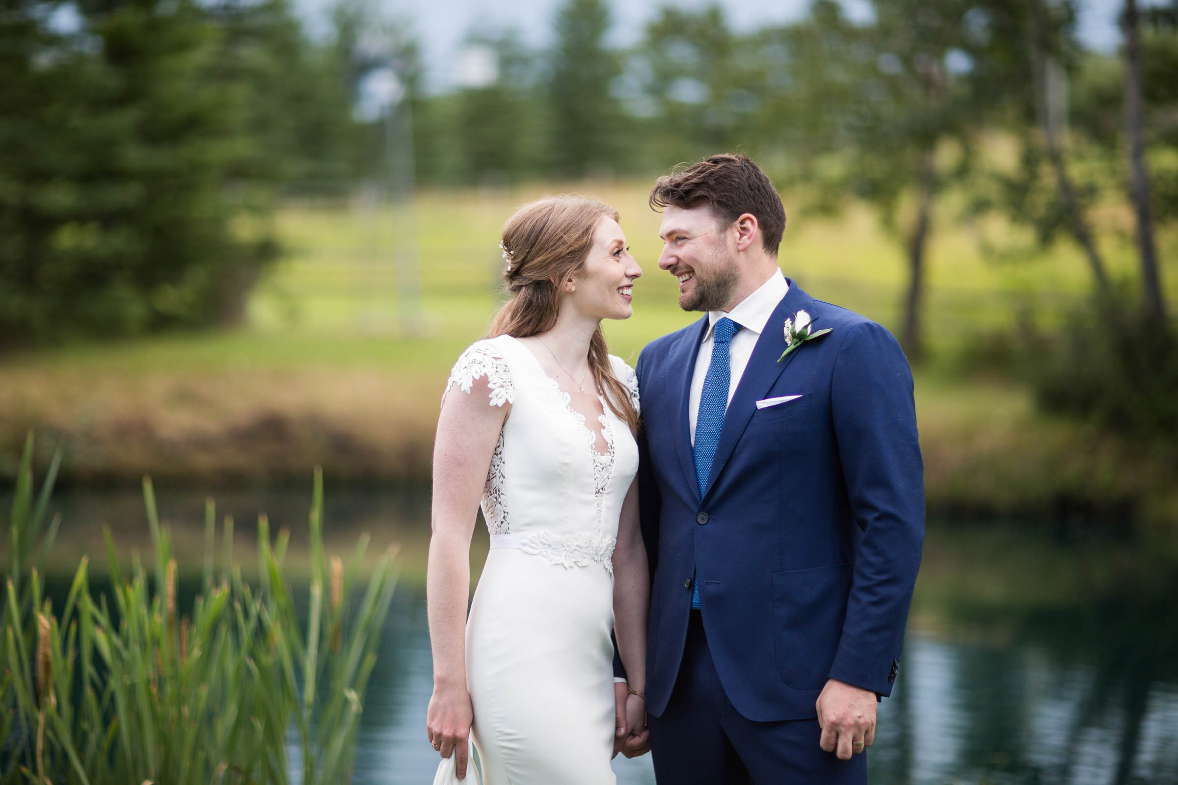 Bride and groom looking into each others eyes in front of small lake and greenery