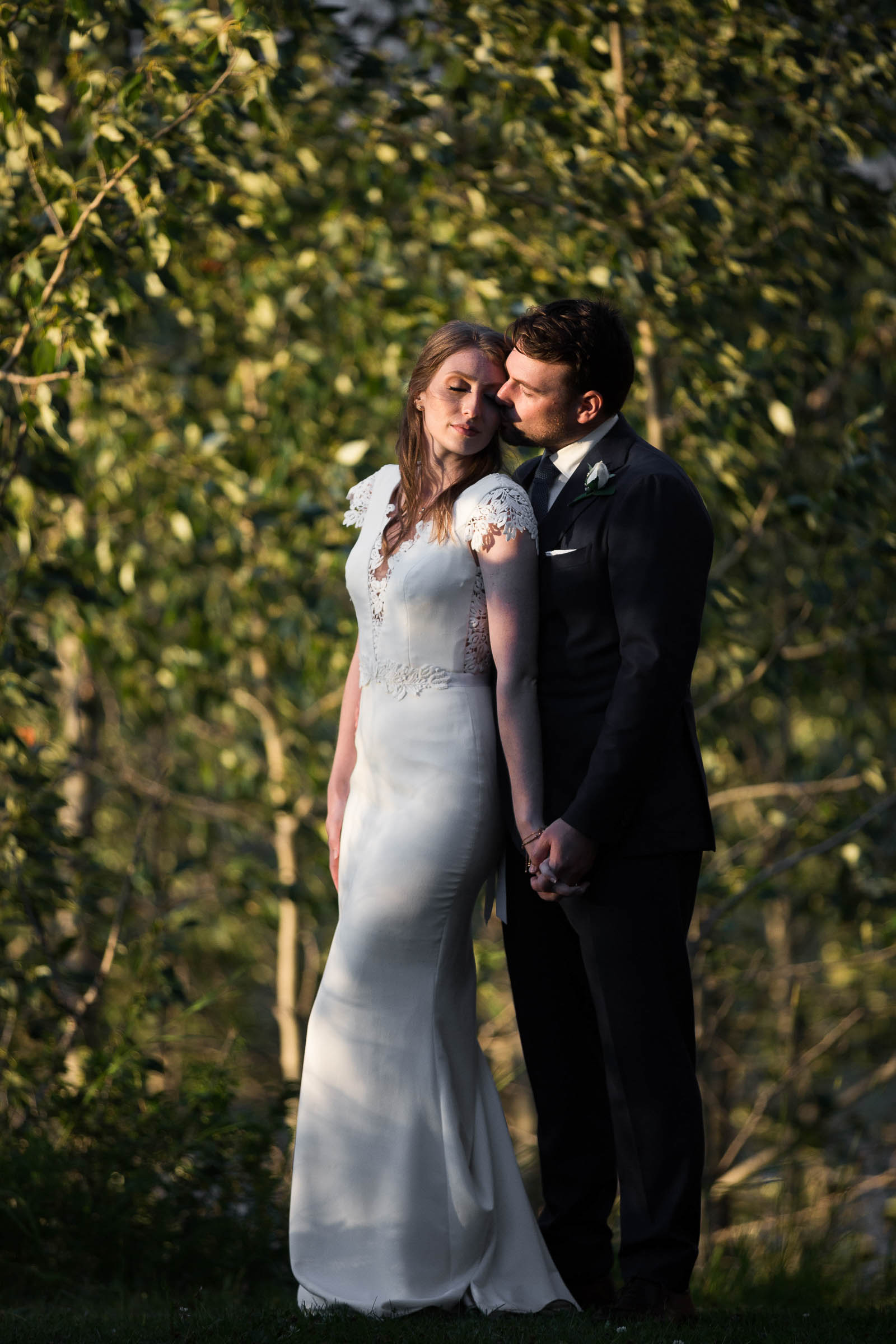 Bride and groom embracing at golden hour in front of wall of greenery