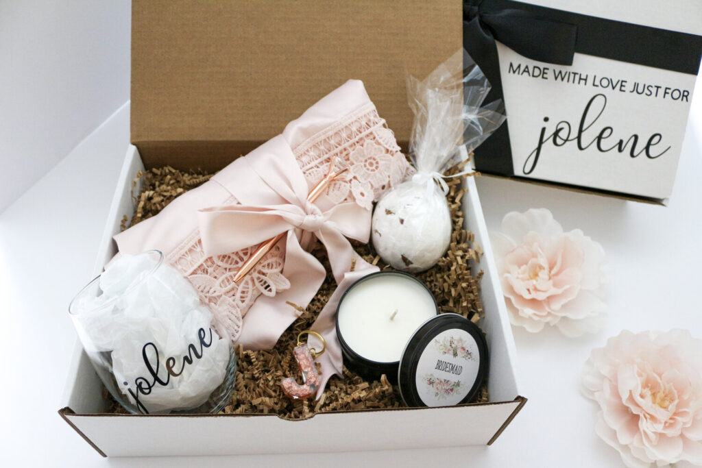 Bridesmaid proposal box with a stemless wine glass, pink robe, bath bomb, candle, and pen in a personalized box with black bow.