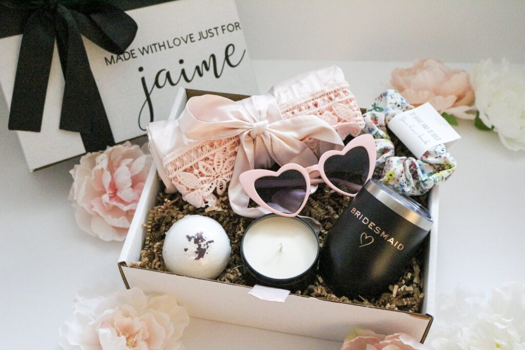 Bridesmaid proposal box with a stemless wine tumbler, pink robe, bath bomb, candle, pink heart sunglasses, and scrunchie in a personalized box with black bow.